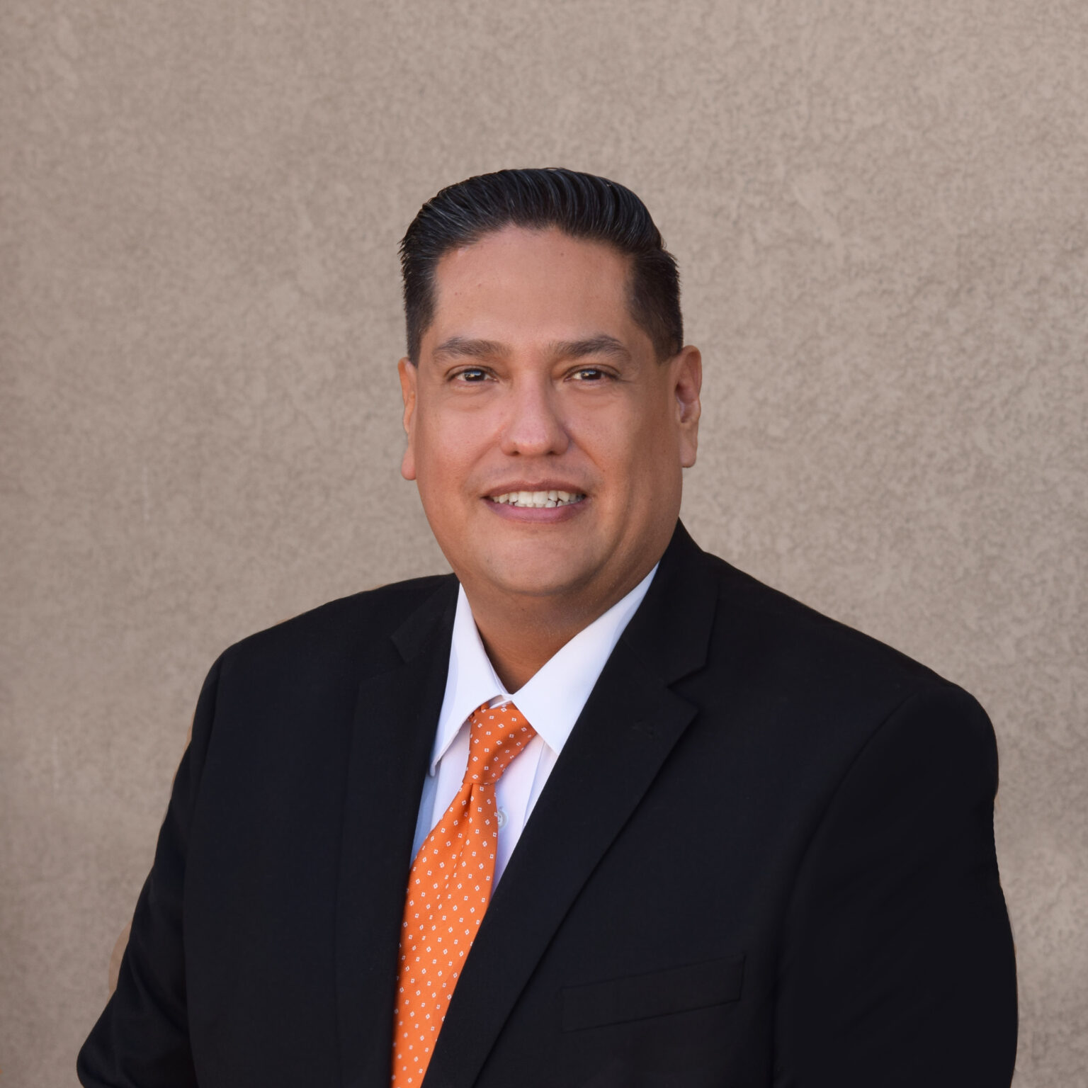 Victor Nava posing for his headshot wearing a black suit, white shirt and orange tie.